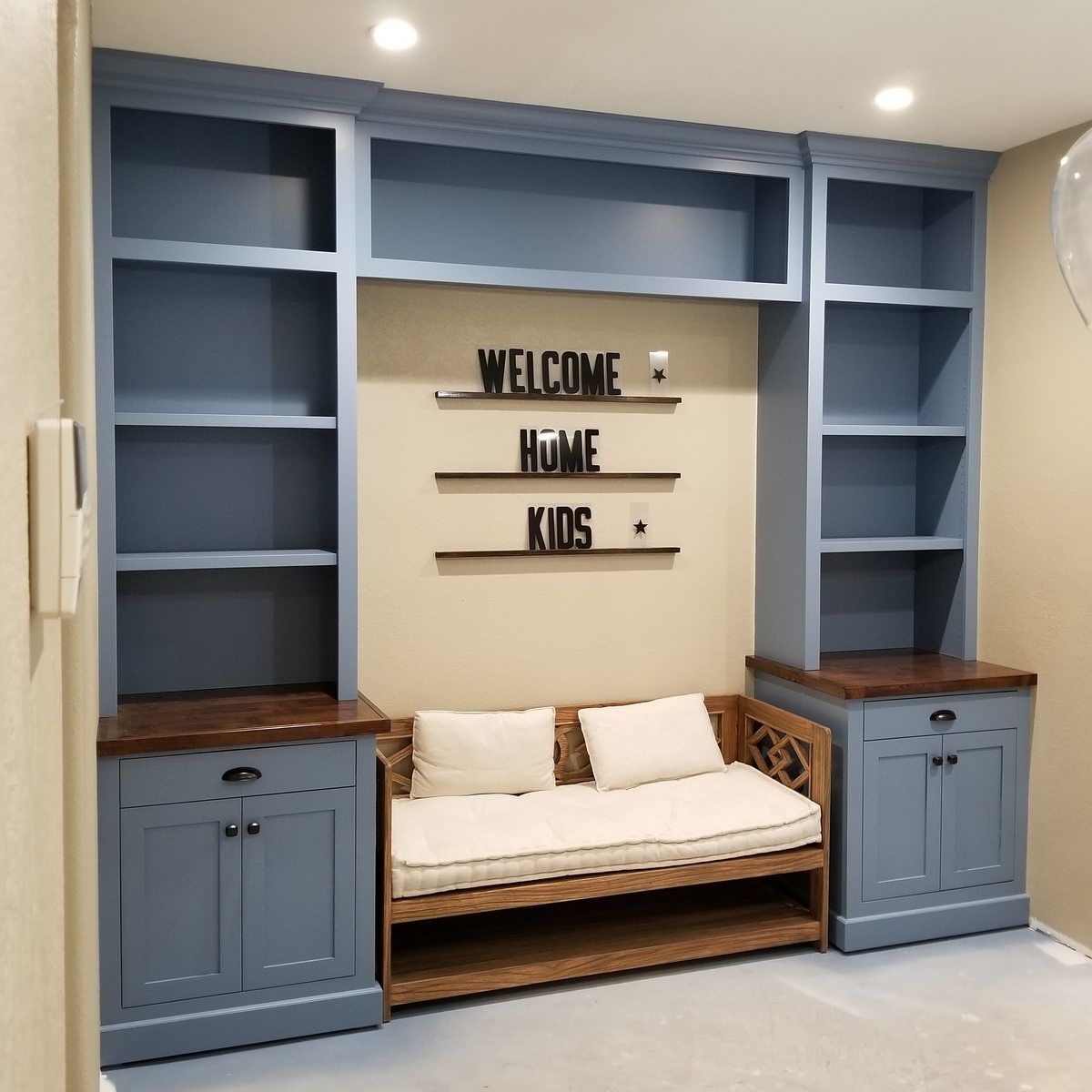 A blue built-in mudroom unit with open shelves on both sides of a bench, under a Welcome Home Kids sign.